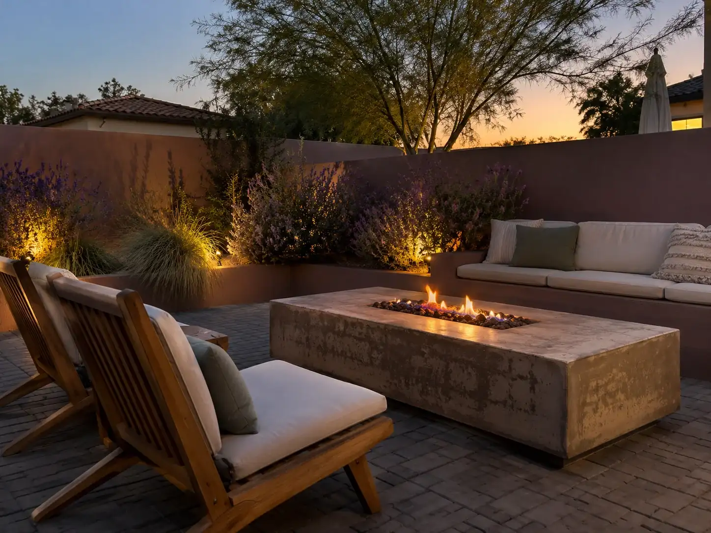 Paver patio at dusk with a rectangular fire table, lounge chairs and lit planting along the wall